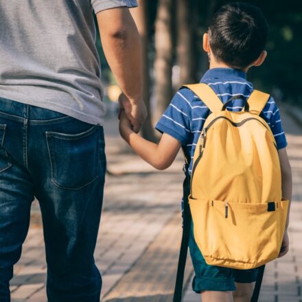 Father walking his son to kindergarten, walking away from camera. Boy has a yellow backpack.