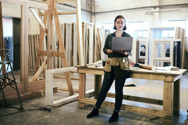 Woman stand in a carpentry shop, wearing a tool belt and holding a Dell computer.