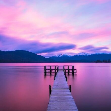Dock leading out into a lake, with calm water. Pink and purples colors in the clouds in the background above the hills, just before sunrise.