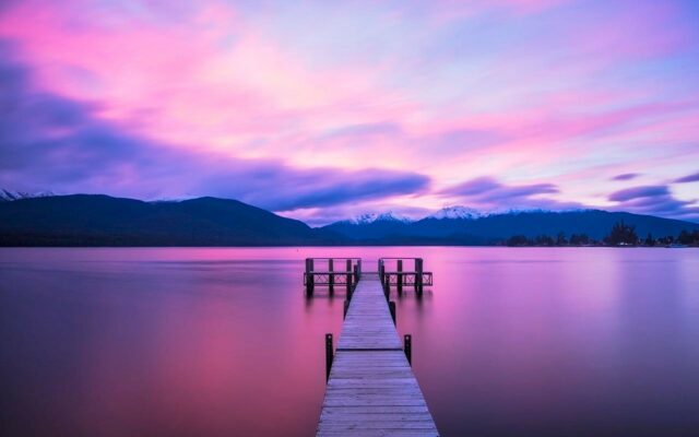 Dock leading out into a lake, with calm water. Pink and purples colors in the clouds in the background above the hills, just before sunrise.