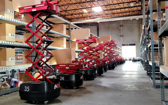 A row of red and black inVia Robotics robots are lined up in a warehouse, with hydraulic lifts extended at various heights.