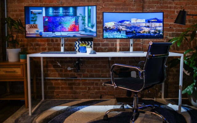 Dell Ultrasharp 40" and 34" monitors positioned side by side on a desk in front of a brick wall with an office chair in the foreground.