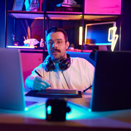 Young attractive man podcaster in glasses explaining concepts with stylus on tablet, in modern home-studio in neon light. Concept of youth people with social media and smart working, lifestyle, online