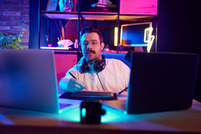 Young attractive man podcaster in glasses explaining concepts with stylus on tablet, in modern home-studio in neon light. Concept of youth people with social media and smart working, lifestyle, online